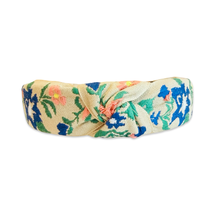 top view of a wide knotted fabric headband with a natural linen-textured base, covered in colorful floral embroidery featuring cobalt blue, green, peach, and yellow botanical motifs. The headband has a center knot detail and smooth cream inner lining.