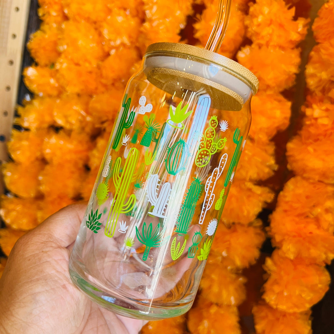 hand holding a glass can tumbler with images of various green cacti and a background of marigold garlands