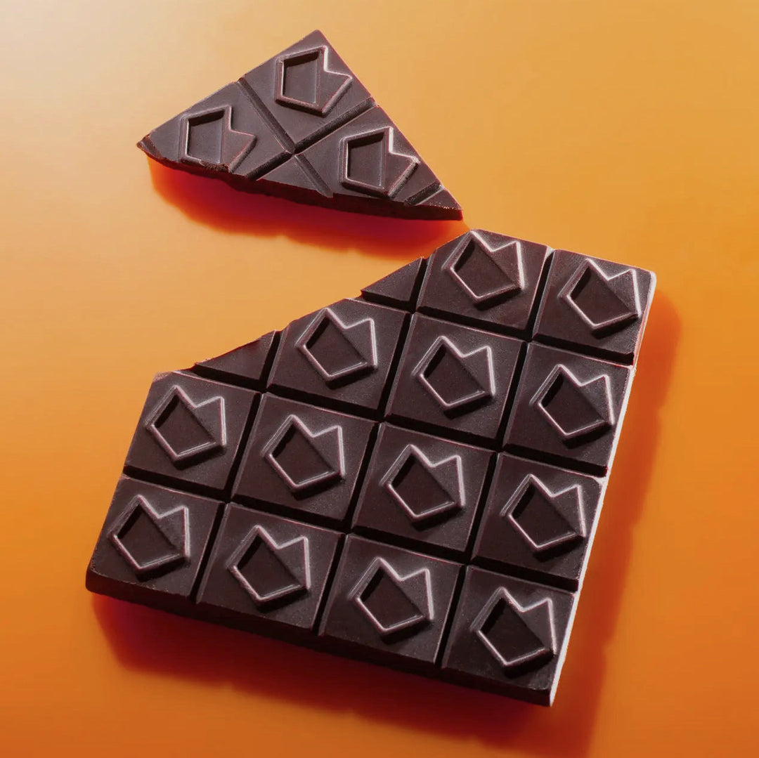 A dark chocolate bar with geometric patterns, partially broken to reveal a sharp triangular piece, set against a warm orange background.