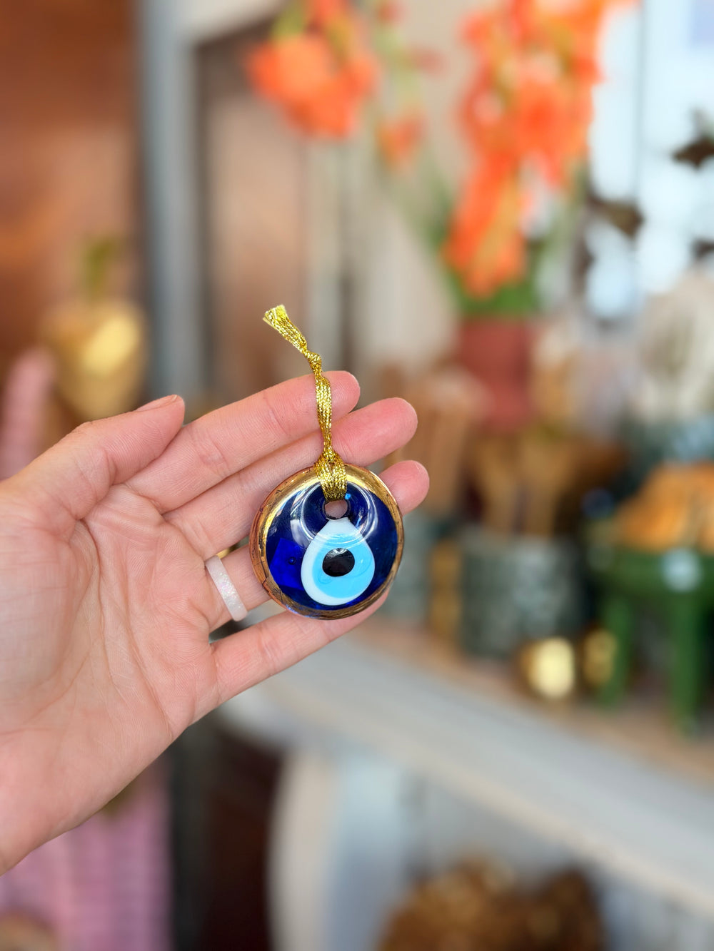 A hand holding a round blue glass evil eye ornament with a gold hanging ribbon, set against a softly blurred indoor background with warm orange flowers.