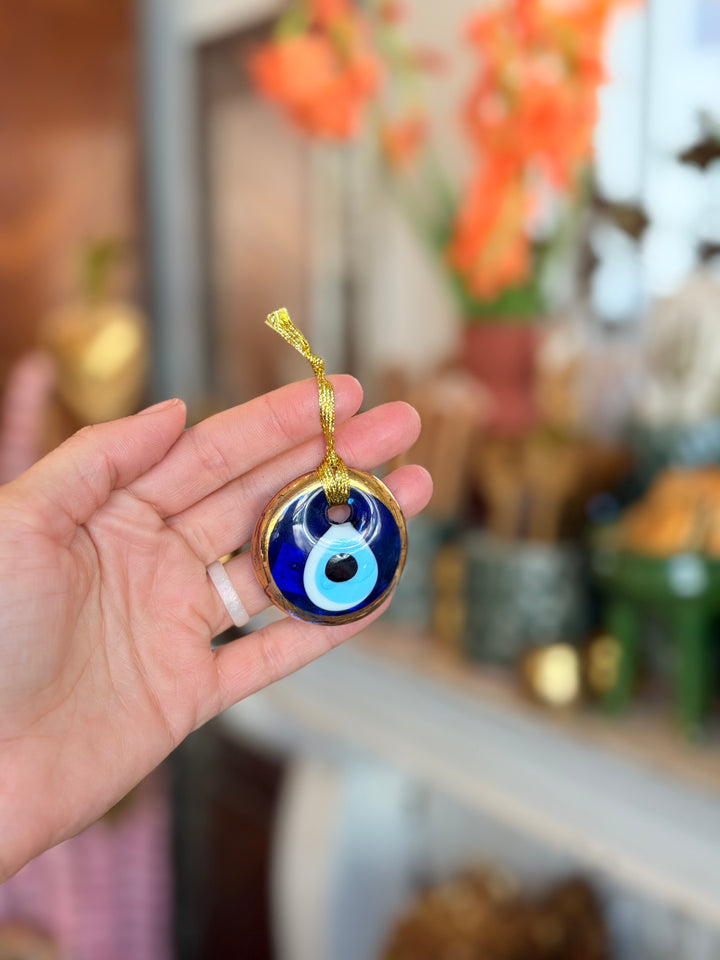A hand holding a round blue glass evil eye ornament with a gold hanging ribbon, set against a softly blurred indoor background with warm orange flowers.