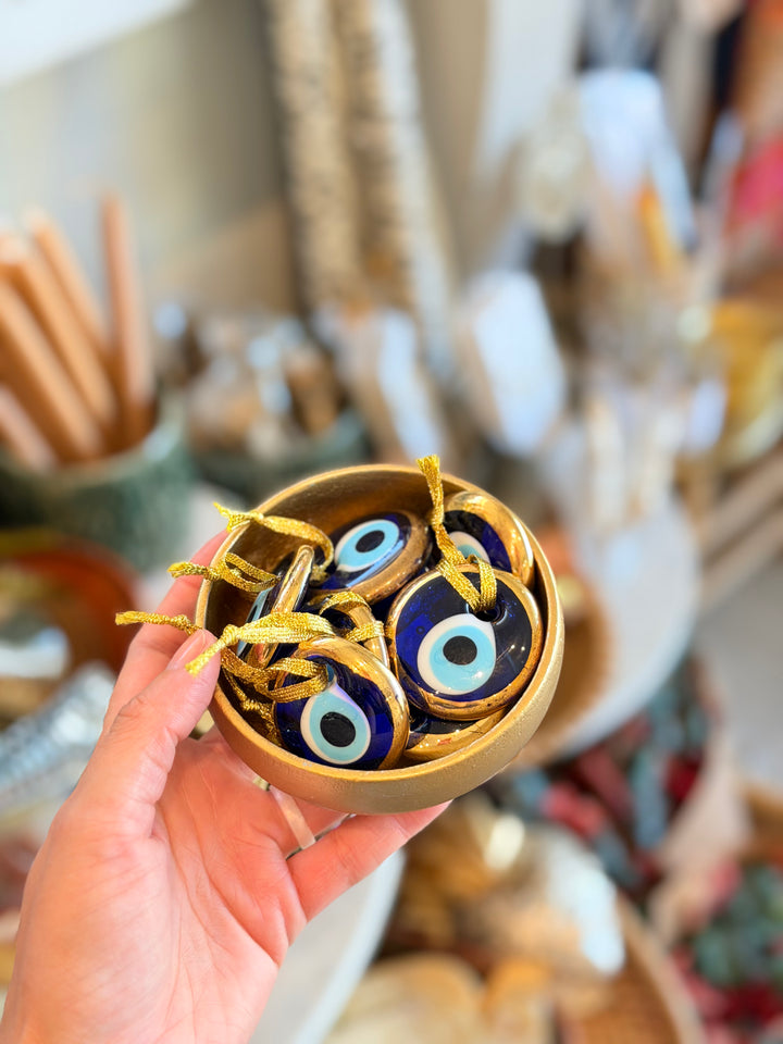 A hand holds a small round gold dish containing several blue and white evil eye glass pendants wrapped in gold ribbon, with a softly blurred shop background.