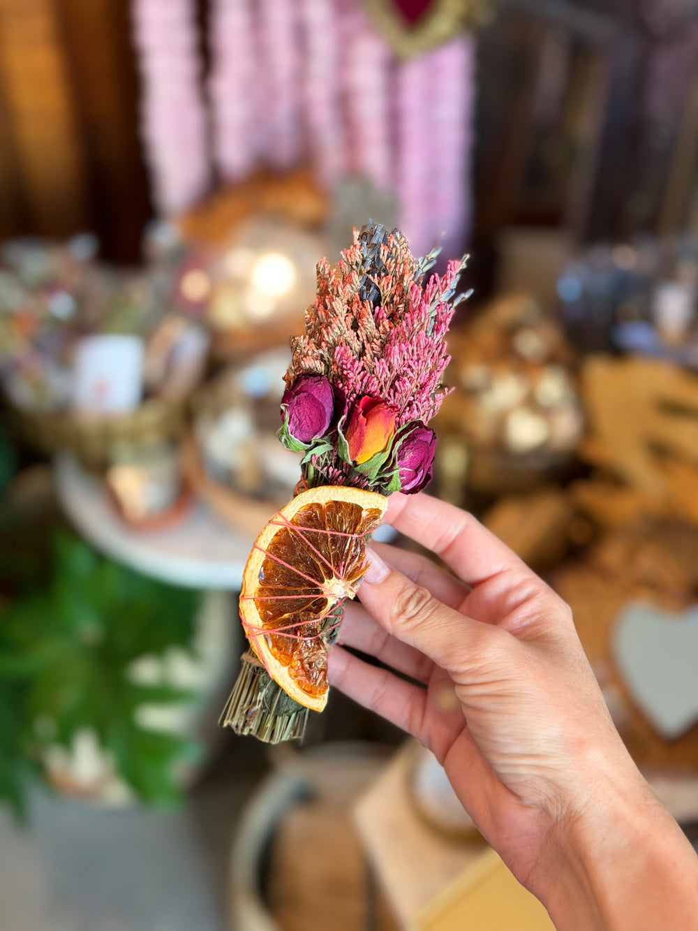 A hand holding a small dried flower bouquet with pink and purple blossoms and a dried grapefruit slice, set against a softly blurred warm-toned background.