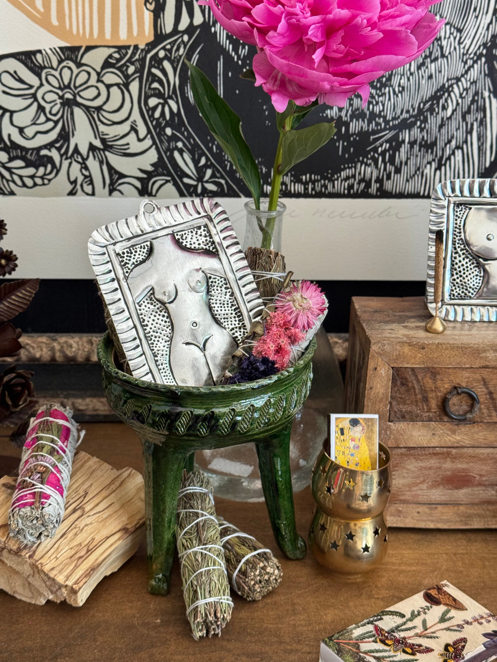 Decorative setting with framed artwork, flowers, and smudge sticks on a wooden surface.
