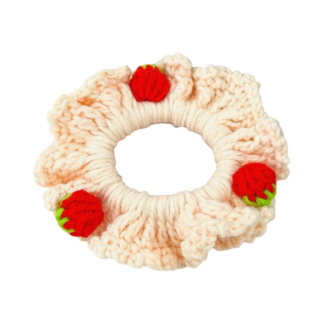 A chunky hand-crocheted hair scrunchie in a soft cream yarn, adorned with three small three-dimensional crocheted strawberries in red and green evenly spaced around the band.