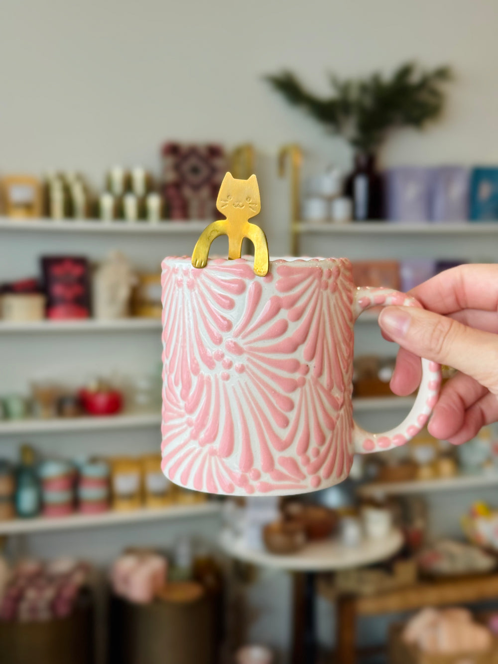 Pink textured mug with a yellow cat-shaped spoon held by a hand in a store setting.