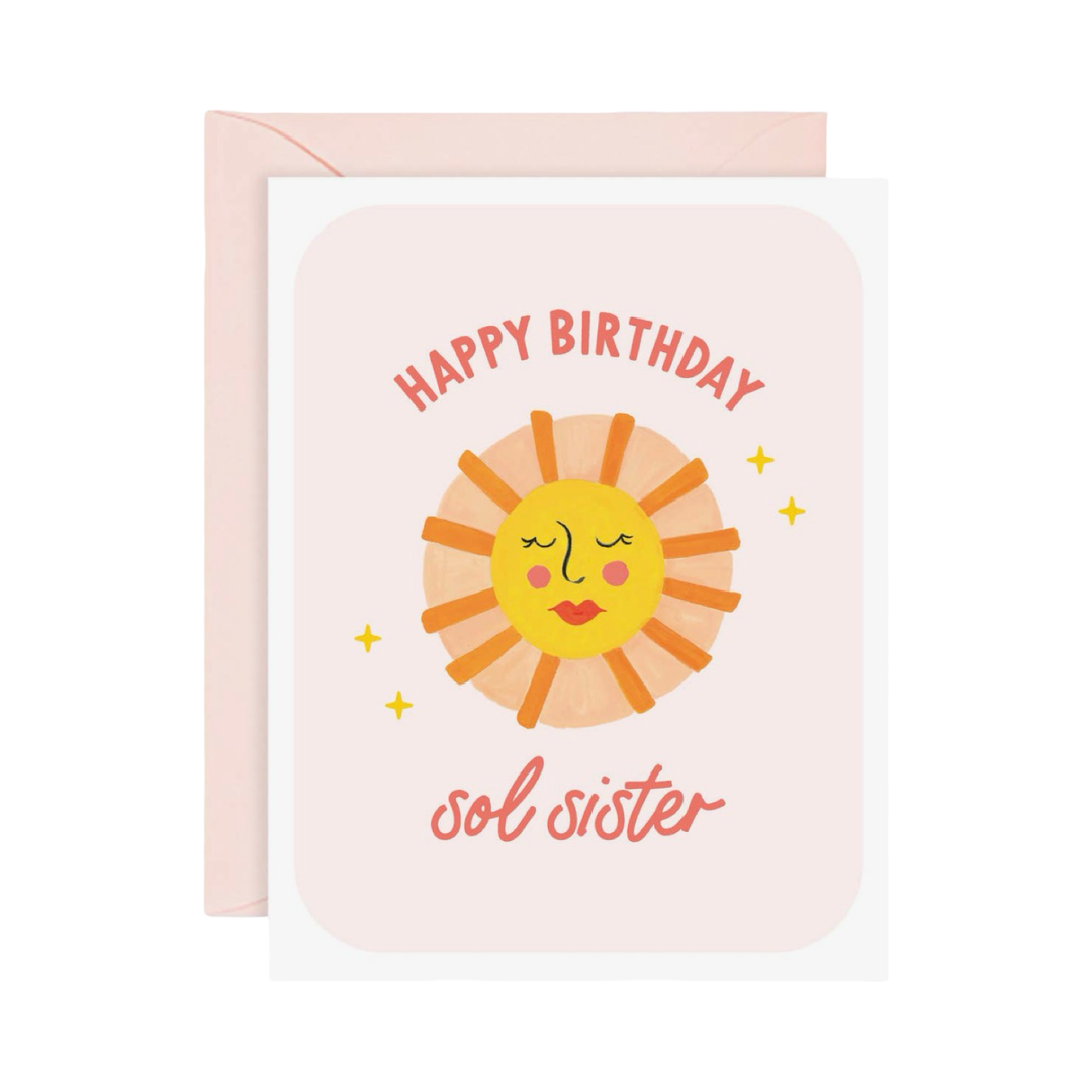 Illustrated birthday greeting card with a blush pink envelope behind it. The card shows a smiling sun face with closed eyes, pink cheeks, and red lips, surrounded by orange sun rays and small gold stars, on a pale pink background. Coral text reads “Happy Birthday” arched above the sun and “sol sister” in cursive script below it.
