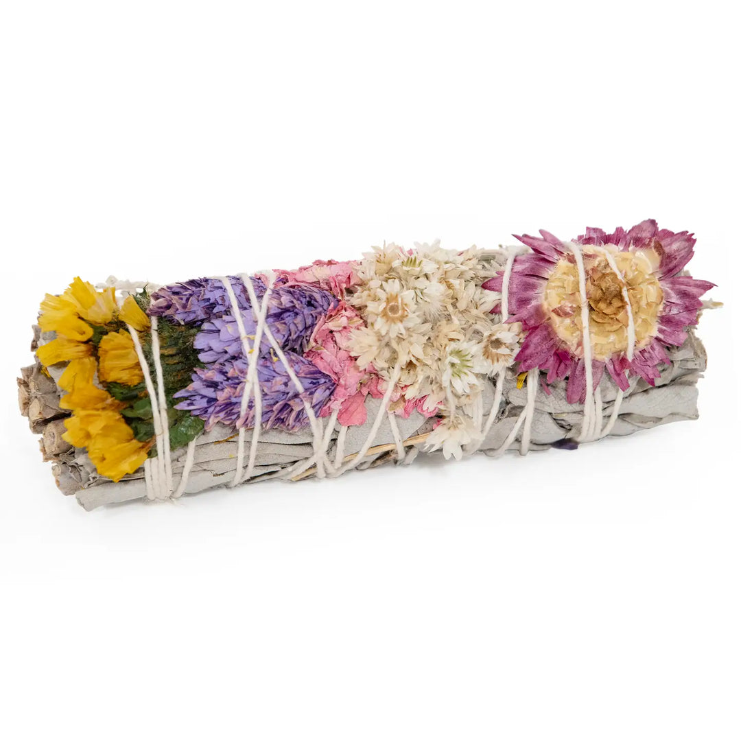 White sage smudge stick wrapped with white string and decorated with bands of yellow, purple, pink, white, and magenta dried flowers.