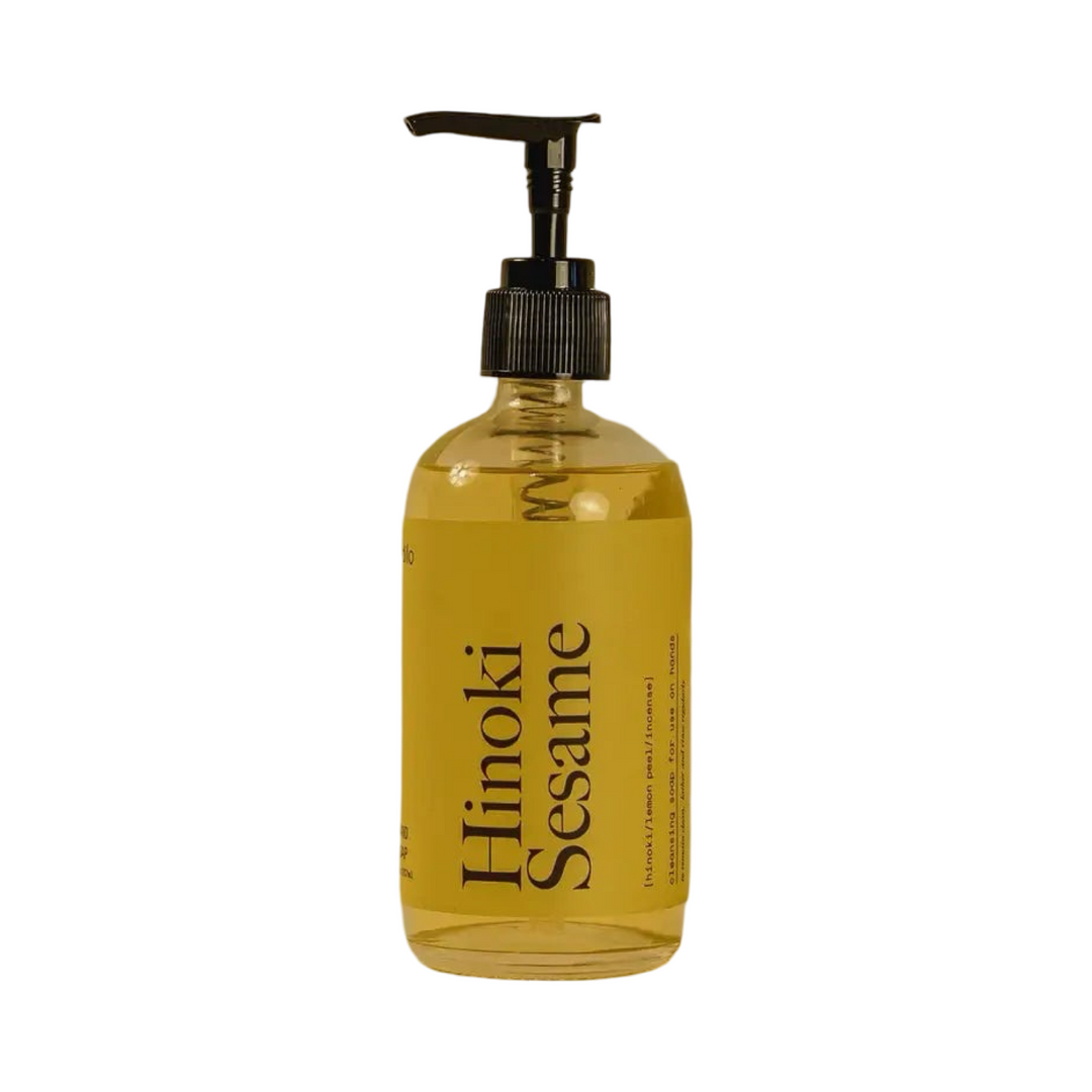 Glass bottle of yellow Hinoki Sesame hand soap with green label and  black pump.