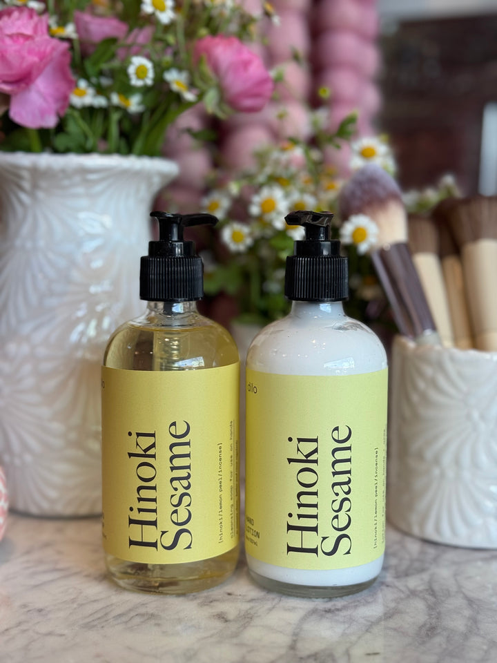 Two bottles of Hinoki Sesame product on a marble surface with flowers and makeup brushes in the background.