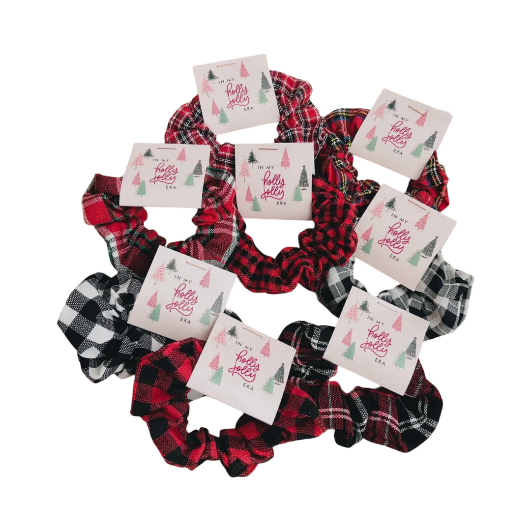 A festive assortment of plaid scrunchies in red, black, and white patterns, each paired with a card that reads “In my holly jolly era” featuring whimsical holiday tree illustrations. 