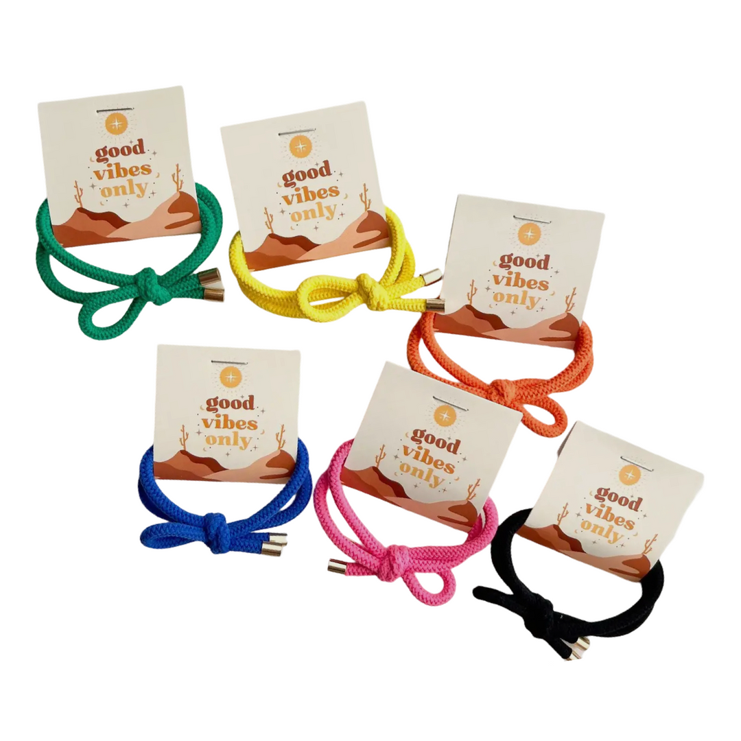 A collection of colorful knotted hair ties in green, yellow, orange, blue, pink, and black, each paired with a card featuring the phrase “Good vibes only” alongside a desert-themed design with cacti and soft earthy tones.