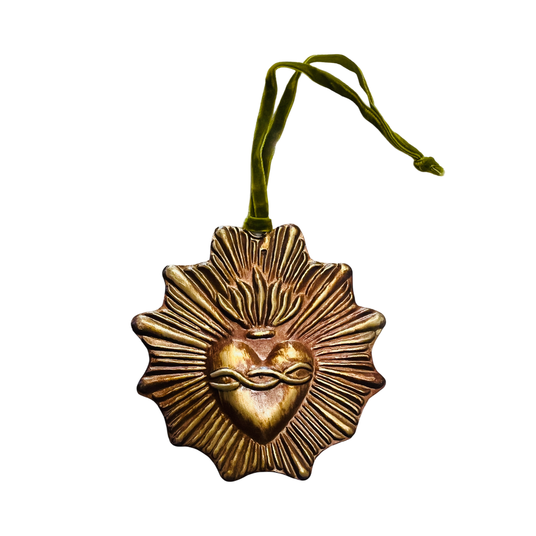 A radiant gold-toned sacred heart ornament with a textured sunburst design surrounding the heart. The heart features intricate detailing, including a crown of thorns and a flame at the top. It is hung by a green velvet ribbon.