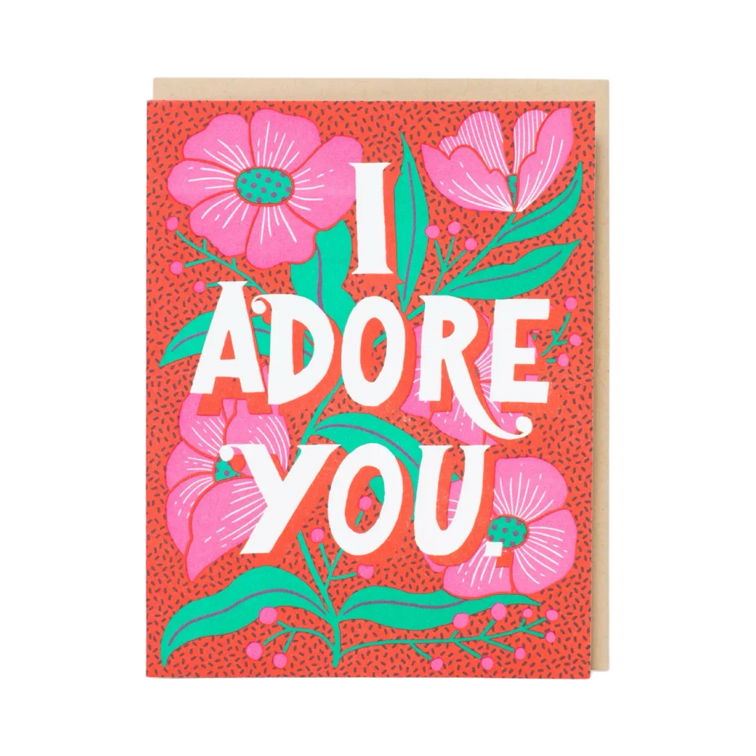 A brightly colored greeting card featuring the bold text "I ADORE YOU" in white, surrounded by vibrant pink flowers, green leaves, and a textured red background. The card is paired with a kraft envelope.