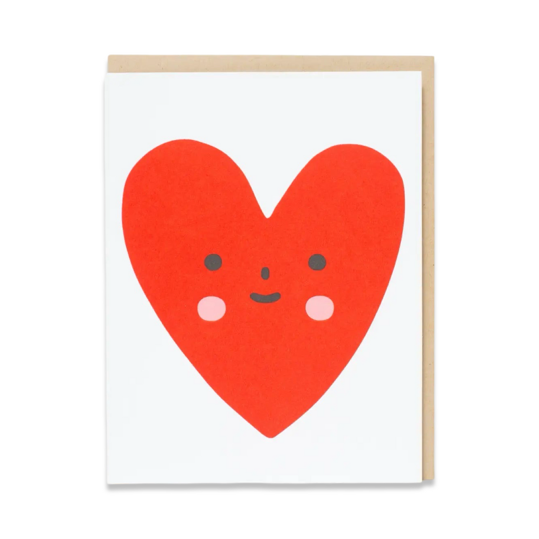 A greeting card with a cute red heart illustration featuring a smiling face with rosy cheeks on a plain white background. Includes a kraft envelope.