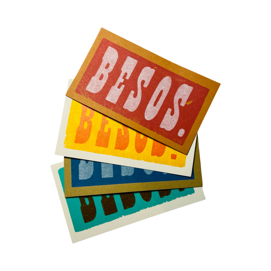 A stack of colorful mini cards with the word "BESOS." printed in bold, vintage-style lettering. The cards come in an assortment of red, yellow, blue, and teal backgrounds, adding a playful and heartfelt touch.