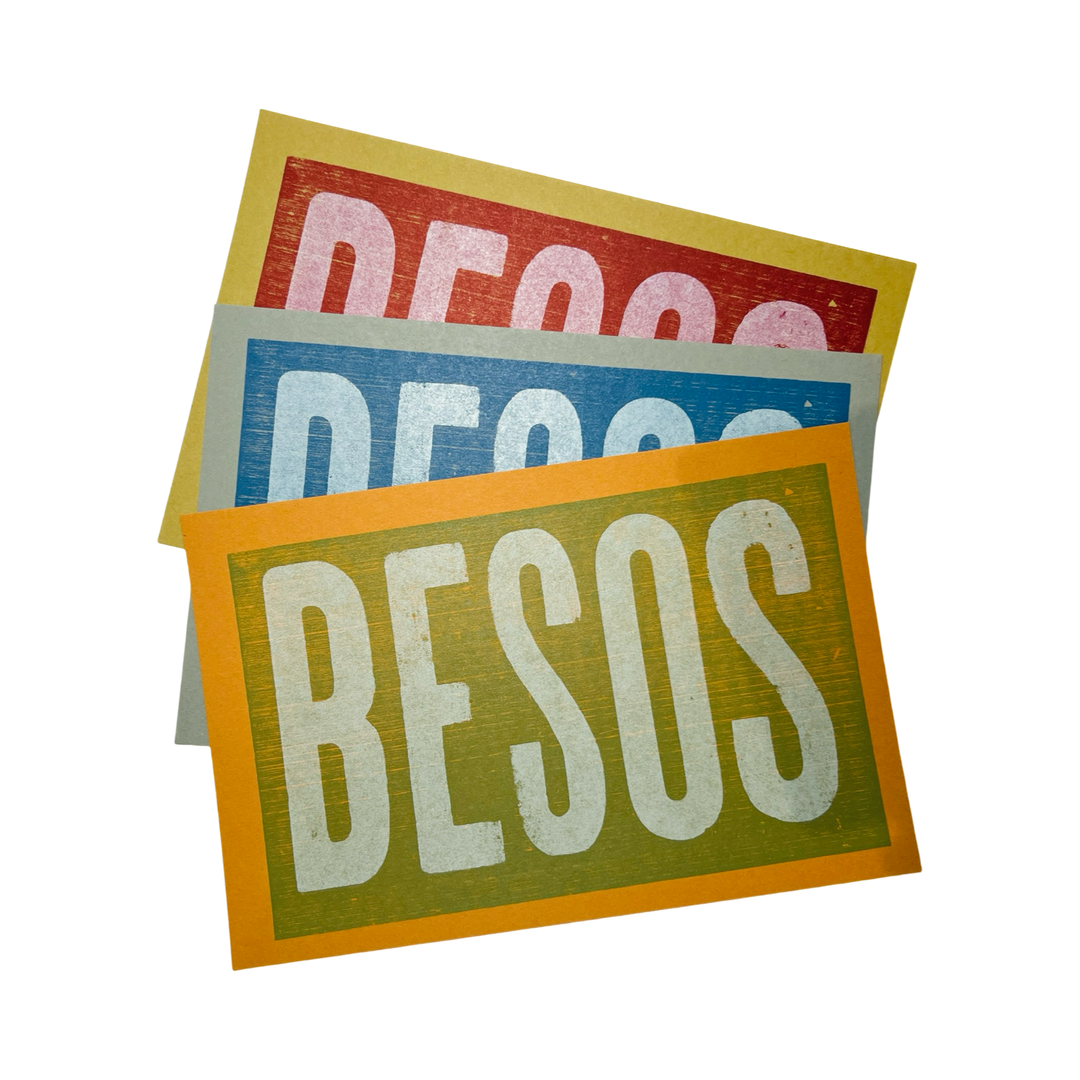 A set of three "BESOS" letterpress prints stacked together, each with a different bold background color—red, blue, and green. The vintage-style typography is prominently displayed, giving the prints a rustic, handcrafted appeal.