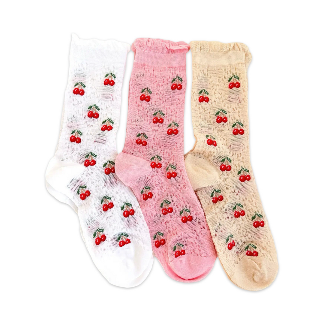 A set of three lace-patterned socks in white, pink, and beige, each decorated with small embroidered red cherries and green stems, laid flat on a neutral fabric background.