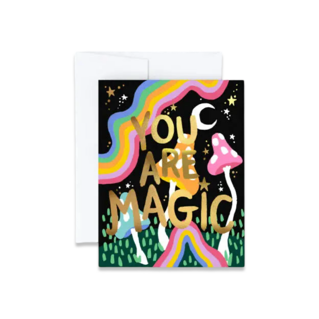 A vibrant greeting card with the phrase "YOU ARE MAGIC" in bold, gold lettering. The design features a whimsical scene with rainbows, stars, a crescent moon, and colorful mushrooms set against a black background. Comes with a white envelope.