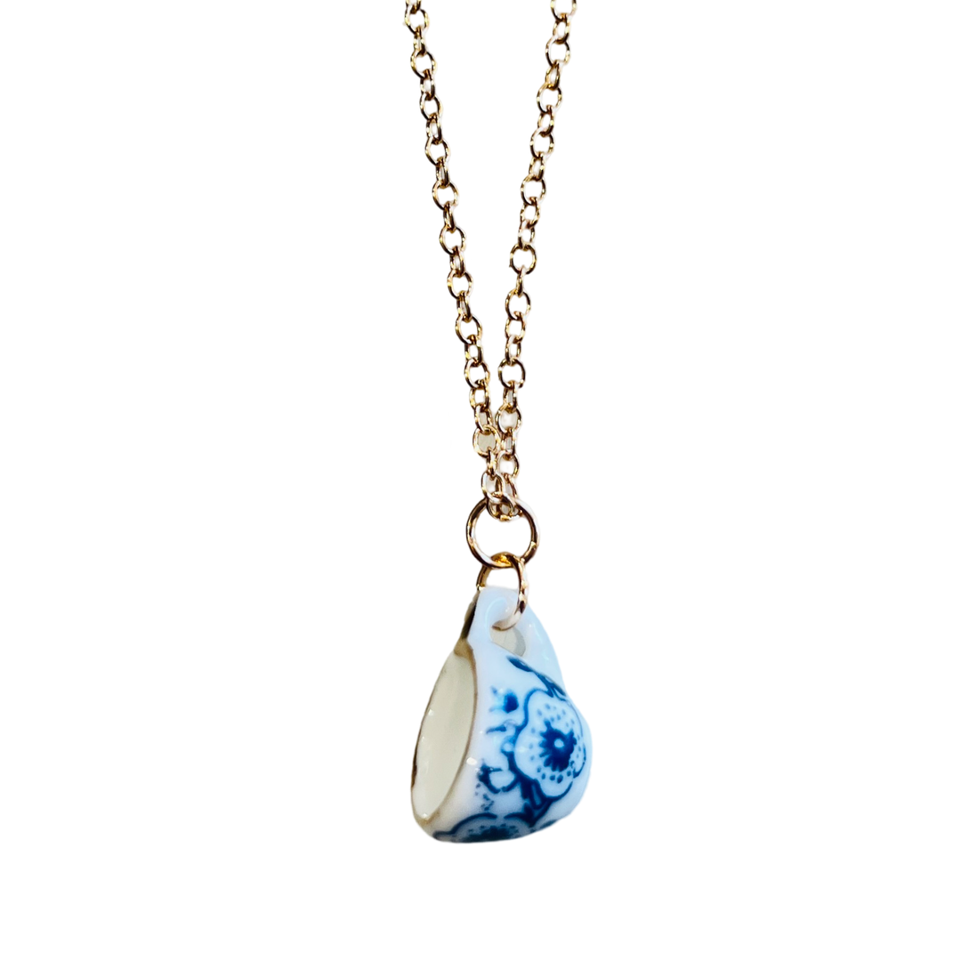 Miniature ceramic tea cup with blue floral designs hanging from a gold-tone chain on a white background.