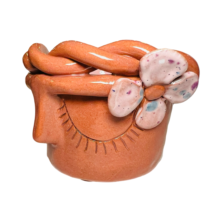 side view of a terracota espresso cup made of barro (clay) with a glazed finish, featuring a face design, pink speckled flower and braided detailing around the rim.