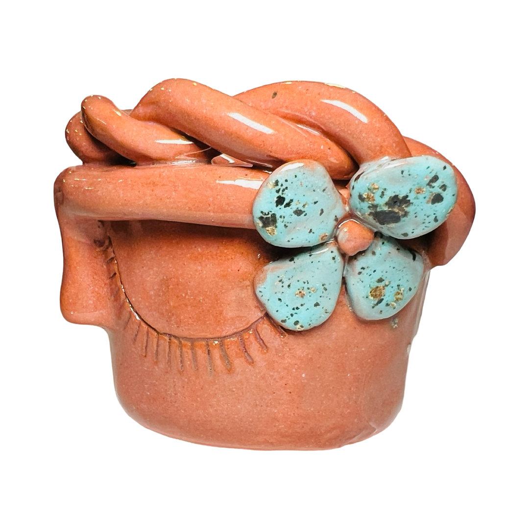 side view of a terracotta espresso cup made of barro (clay) with a glazed finish, featuring a face design, teal speckled flower and braided detailing around the rim.