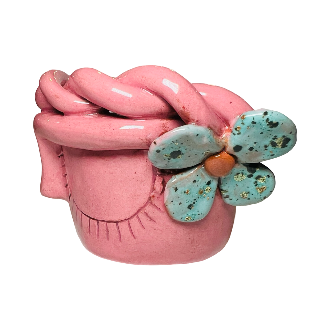 side view of a pink espresso cup made of barro (clay) with a glazed finish, featuring a face design, speckled teal flower and braided detailing around the rim.