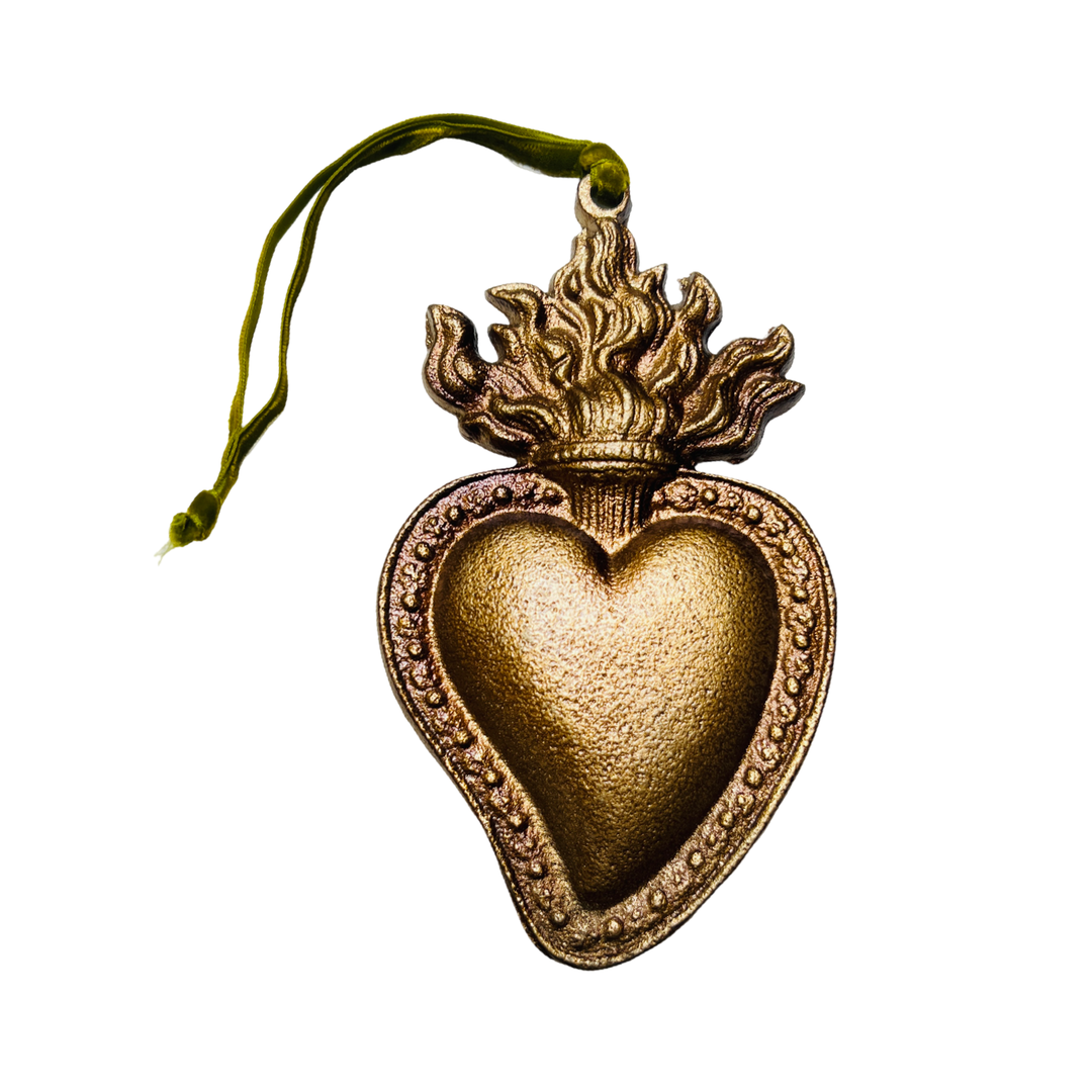 Gold flaming sacred heart ornament with intricate details and a green ribbon for hanging. The heart is surrounded by a textured border and topped with flames.