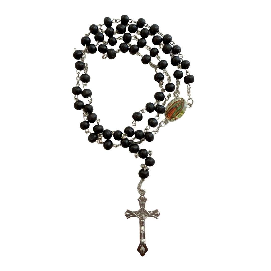 Black wooden bead rosary with silver-tone crucifix and an oval Our Lady of Guadalupe centerpiece, coiled on a white background.