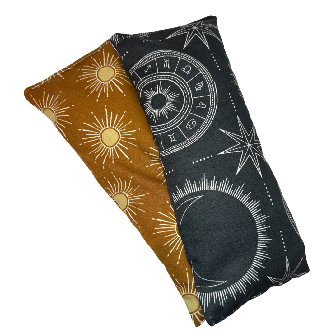 two rectangle eye pillows with one having a golden sun design and the other having a black astrology design