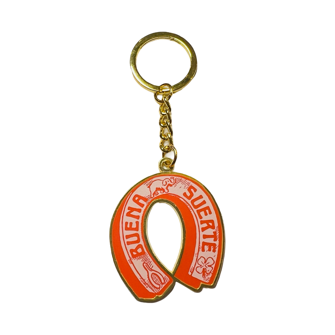 coral and light pink enamel keychain featuring a horseshoe shape, images of an elephant, wish bone, four-leaf clover and the phrase Buena Suerte in coral lettering