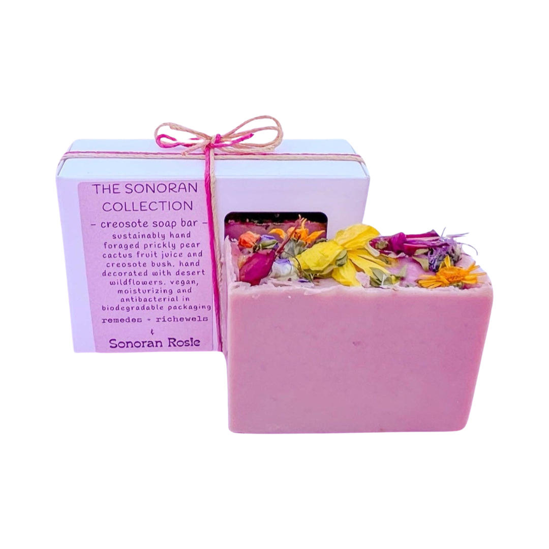 bar of creosote soap featuring dried flowers in front of a white box of soap with a pink branded label