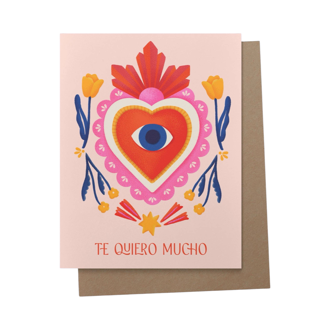 a brown envelope with a blush card and an illustration of a red and pink milagro heart surrounded by orange flowers and a star with an eye in the center of the heart featuring the phrase Te Quiero Mucho in red lettering