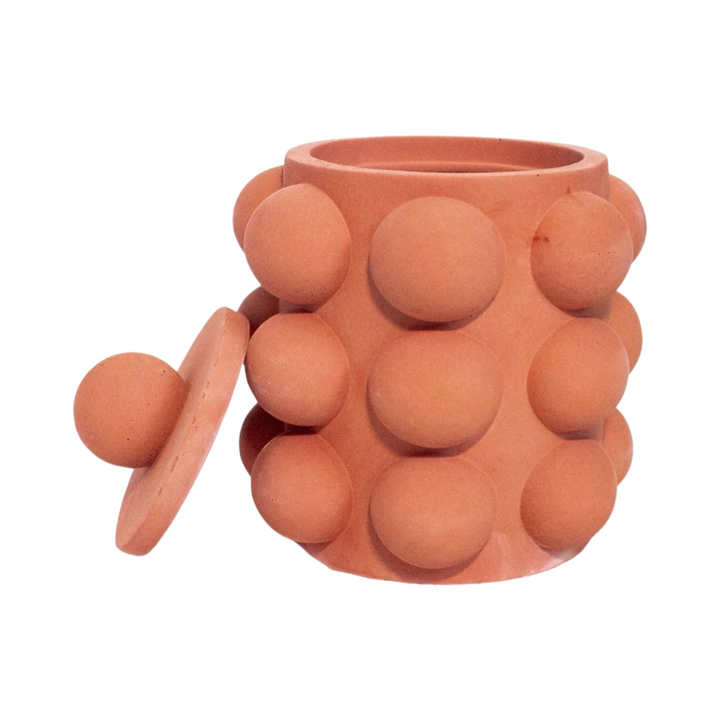 Terracota cylinder shaped candle vessel with a bubble texture and a lid leaning on the side of vessel.