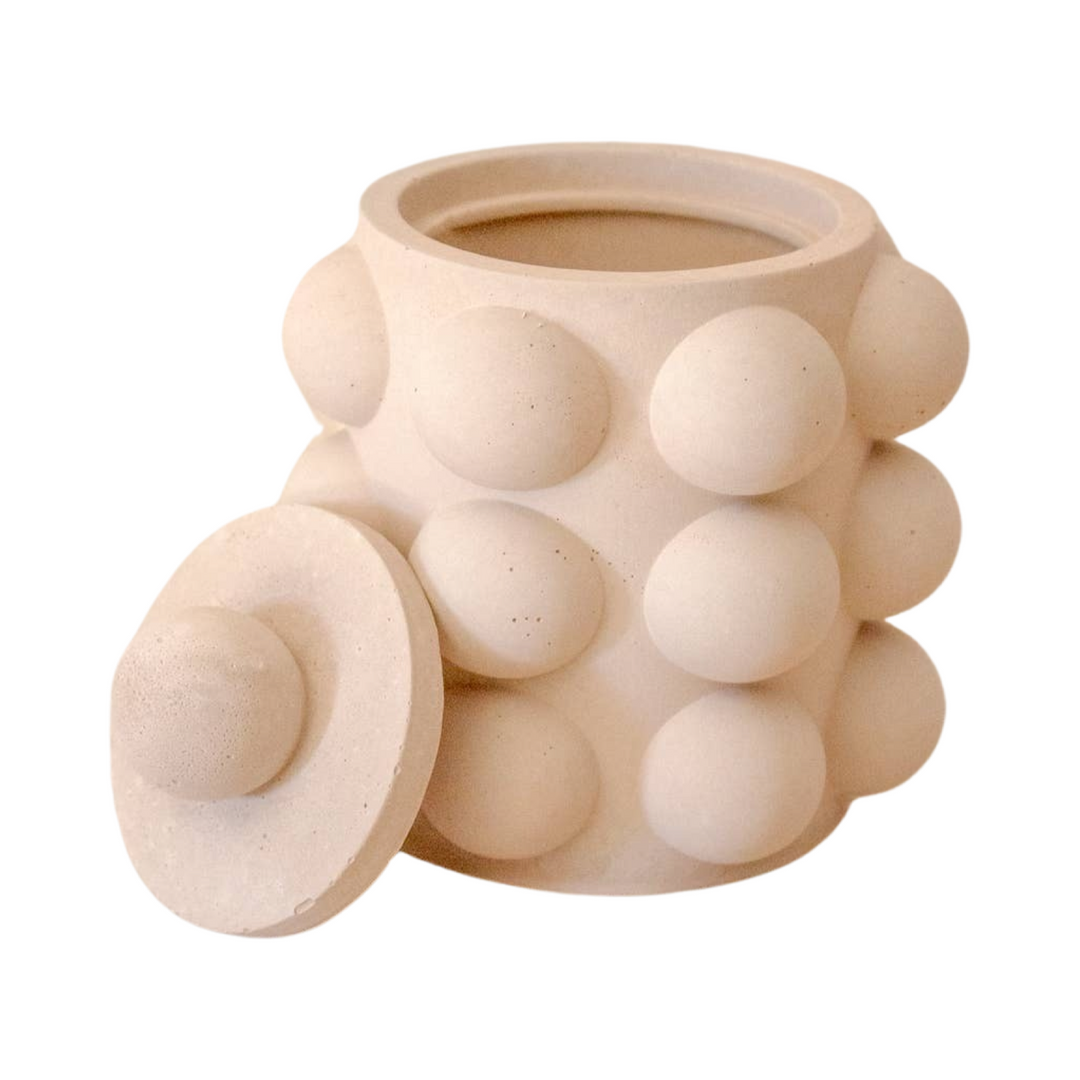 Beige cylinder shaped candle vessel with a bubble texture and a lid leaning on the side of vessel.