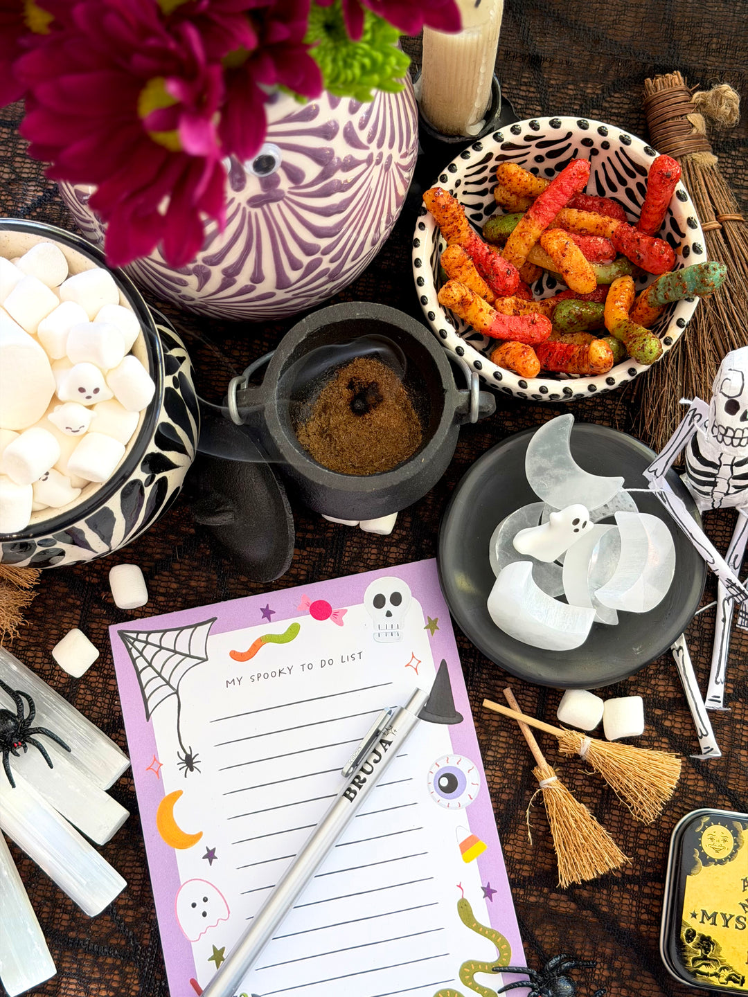 A styled Halloween tabletop scene with a purple 'My Spooky To Do List' notepad, a silver pen, marshmallows, candy corn, a small cauldron, broomsticks, spider decorations, and a patterned vase with flowers on a dark wooden surface.