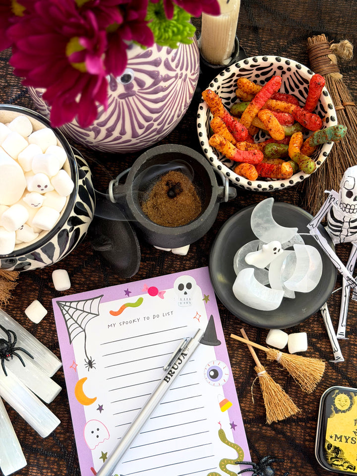 A styled Halloween tabletop scene with a purple 'My Spooky To Do List' notepad, a silver pen, marshmallows, candy corn, a small cauldron, broomsticks, spider decorations, and a patterned vase with flowers on a dark wooden surface.