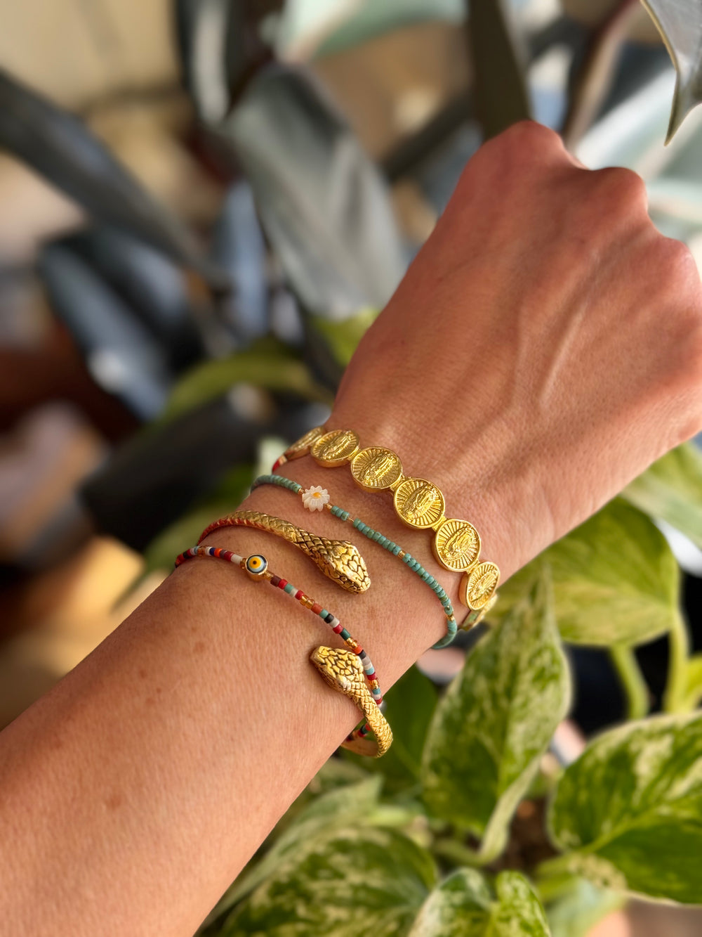 Close-up of a wrist wearing multiple gold and colorful bracelets with a blurred plant background