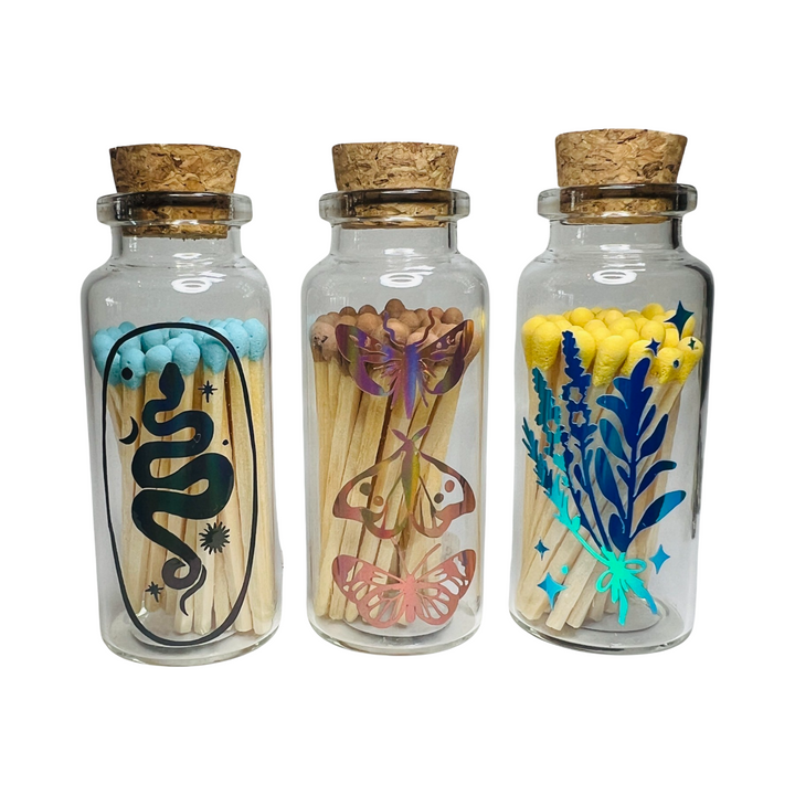 3 clear jars with corks filled with matches featuring holographic images of a snake, butterflies and herb bundle in various colors.