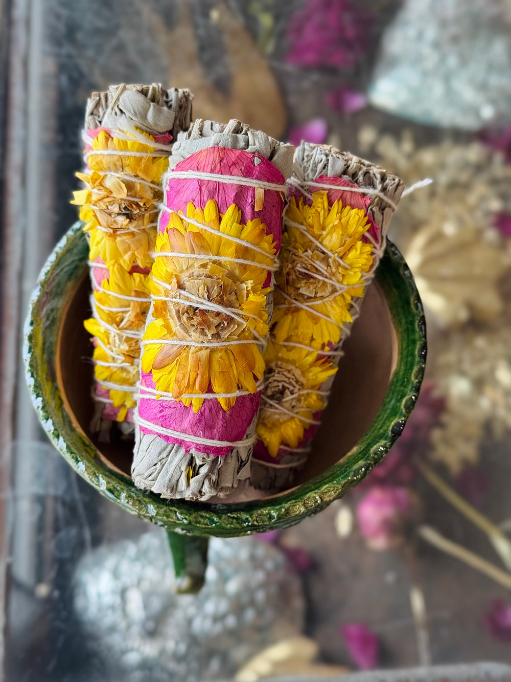 Bundles of white sage wrapped with pink rose petals and yellow sunflowers, tied together with natural string displayed in a green bowl sitting on a dried botanical background.