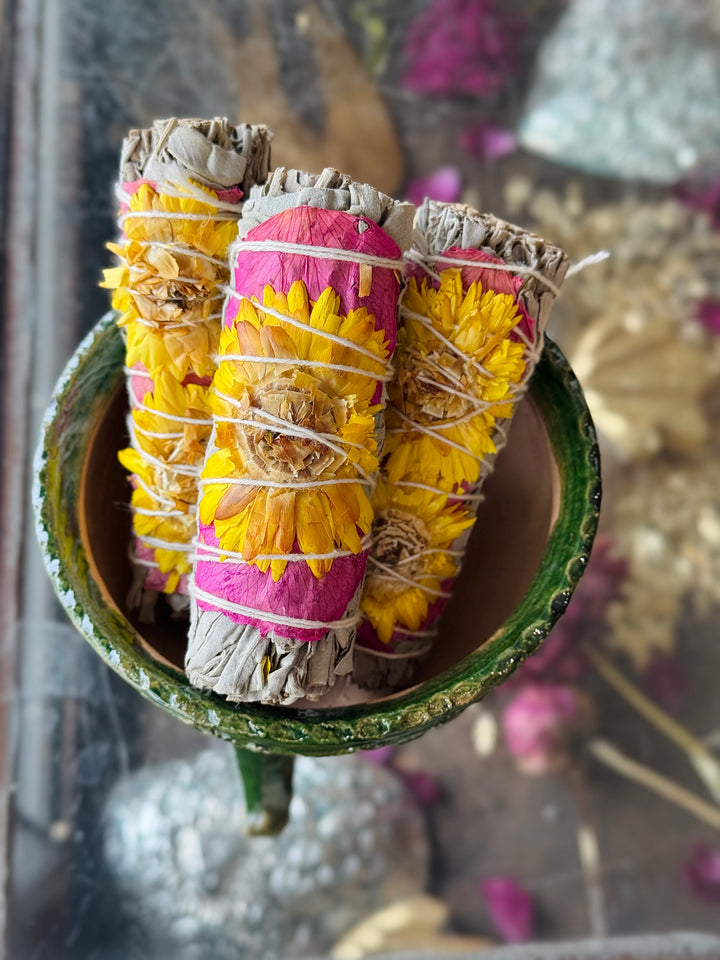 Bundles of white sage wrapped with pink rose petals and yellow sunflowers, tied together with natural string displayed in a green bowl sitting on a dried botanical background.