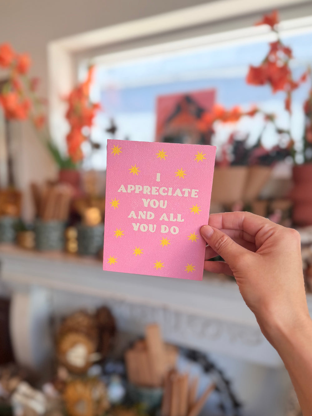 A hand holds a pink greeting card with yellow stars and the text 'I APPRECIATE YOU AND ALL YOU DO' in a brightly lit, blurred indoor setting.