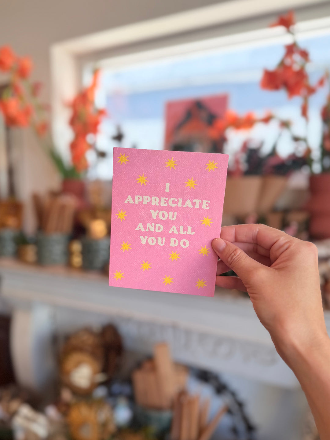 A hand holds a pink greeting card with yellow stars and the text 'I APPRECIATE YOU AND ALL YOU DO' in a brightly lit, blurred indoor setting.