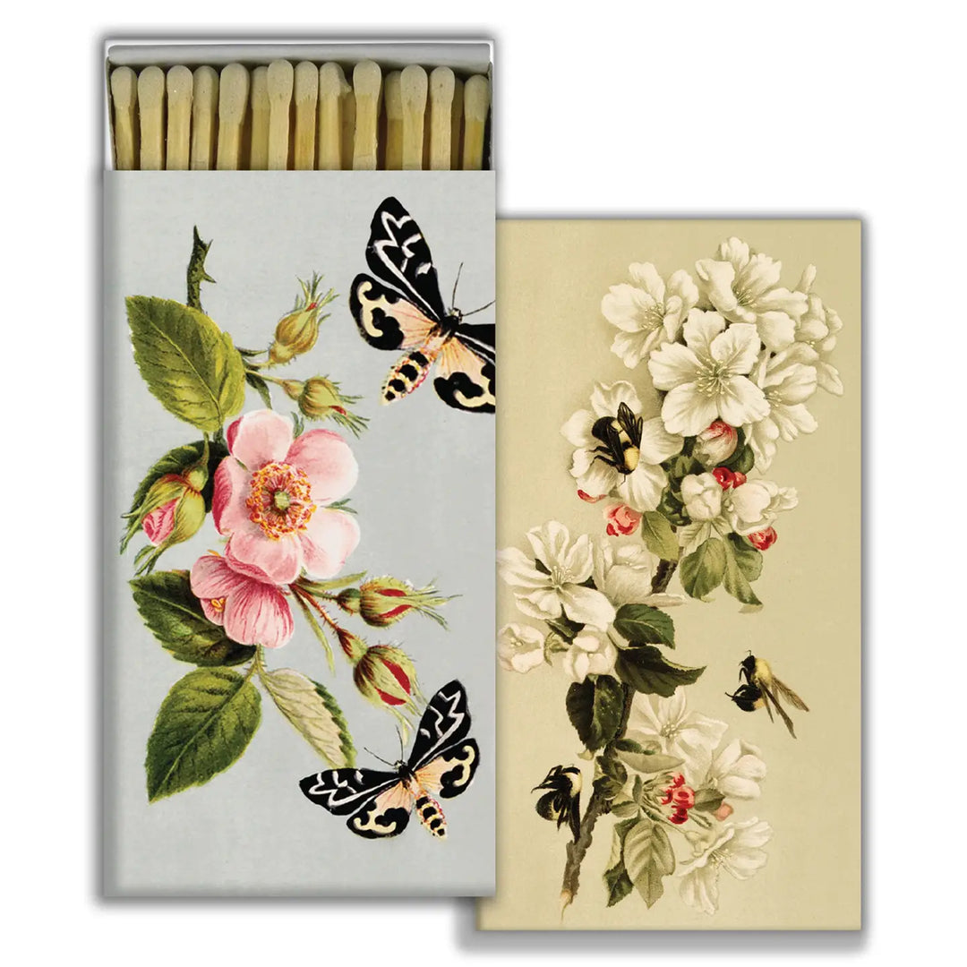 Two rectangular matchboxes with vintage botanical illustrations. One features pink flowers, green leaves, and two black-and-yellow moths on a pale blue background. The other shows white blossoms and bees on a soft beige background. Both boxes contain long, cream-colored matches.
