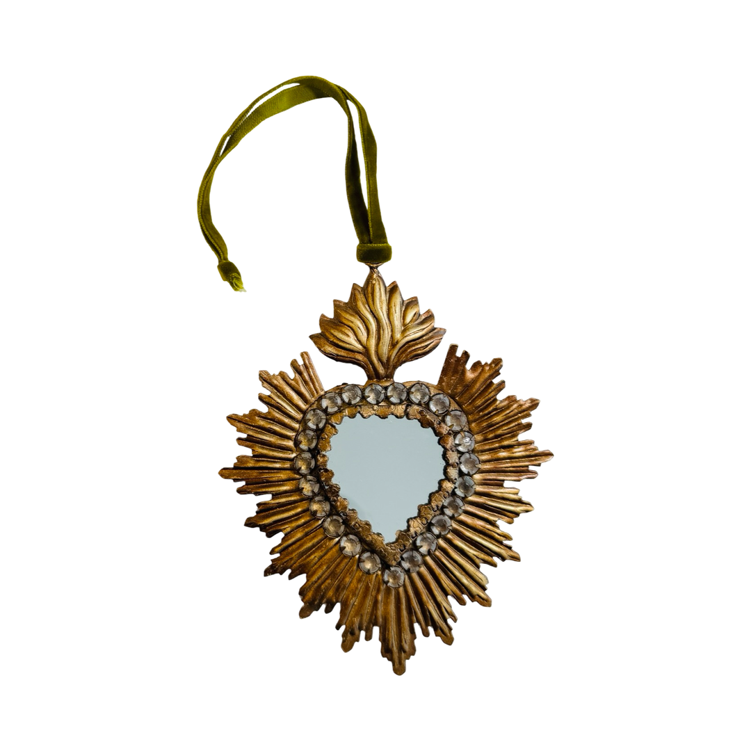A gilded metal sacred heart ornament with a heart-shaped mirror center rimmed with rhinestones, a radiating sunburst, and a flame topper, hung from an olive green velvet ribbon.