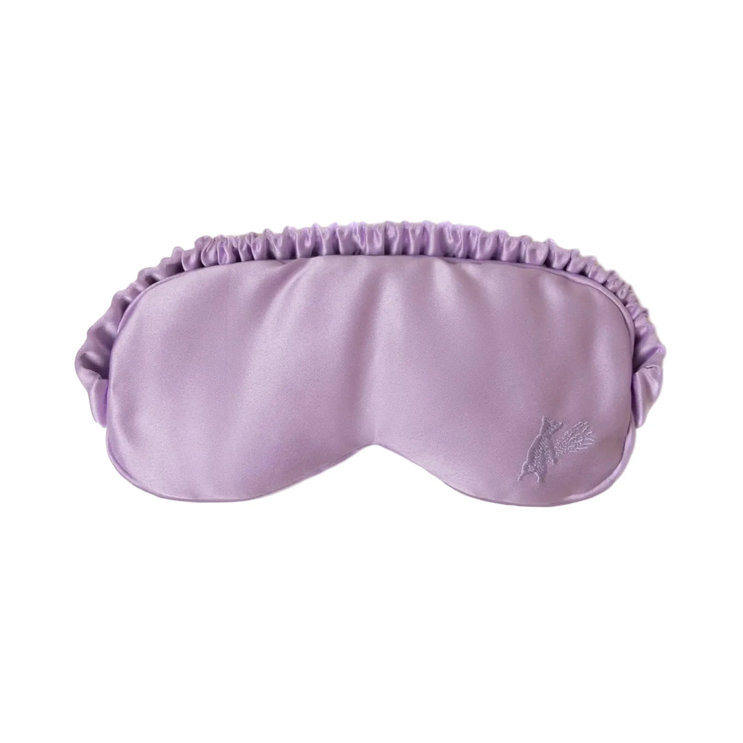A soft lavender satin sleep eye mask with a ruched elasticated band and a small embroidered floral motif