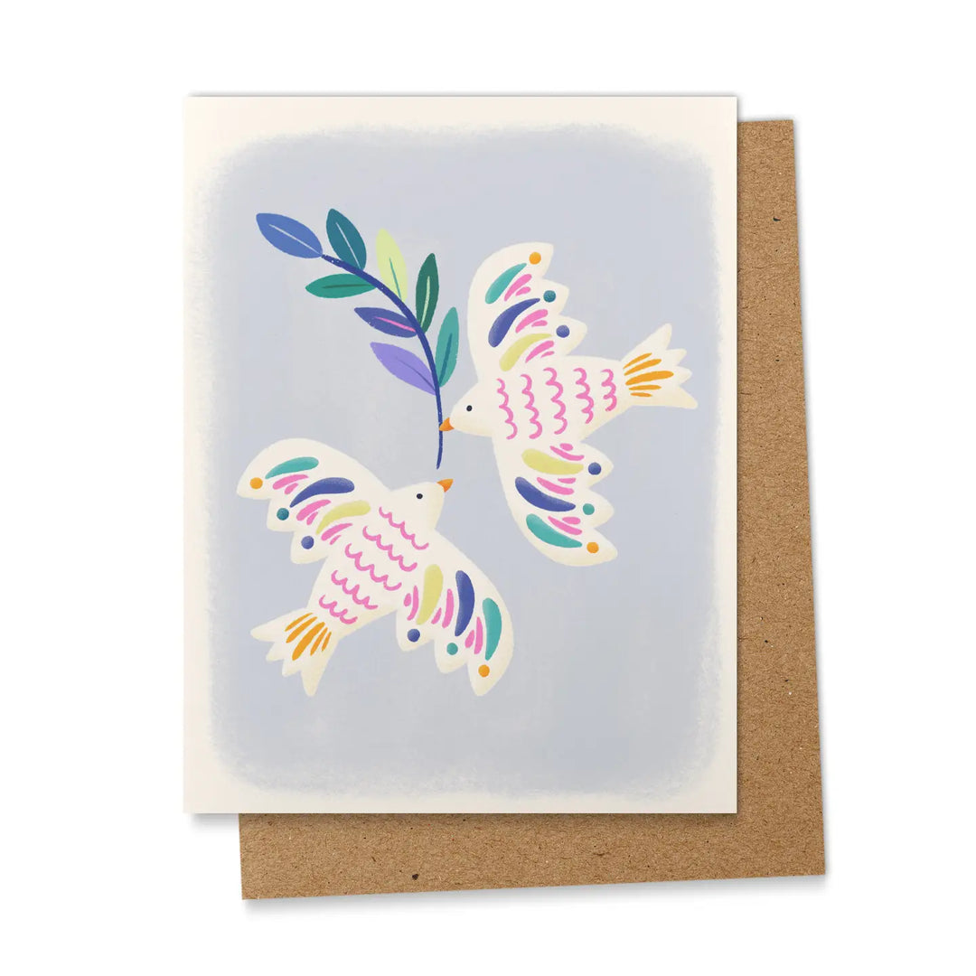 A greeting card featuring two illustrated folk art-style doves in flight, decorated with colorful pink, teal, purple, and yellow details, with one dove carrying a multicolored olive branch, set against a soft periwinkle blue background, displayed propped against a kraft brown envelope.