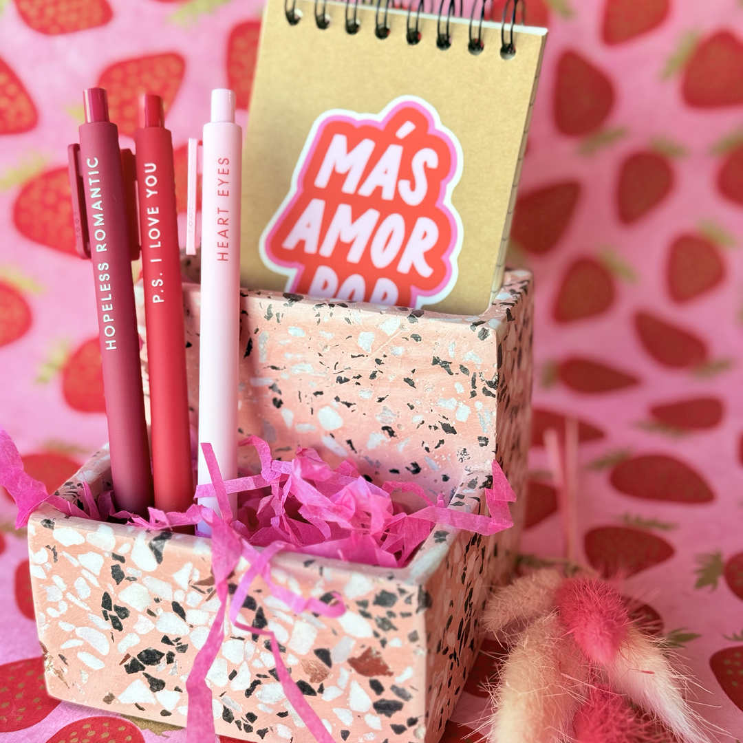 Photo of a pink terrazzo tabletop organizer  with 2 compartments. Also includes notepad with cover art reading "Más Amor Por Favor" & 3 gel pens in shades of pink & red. 