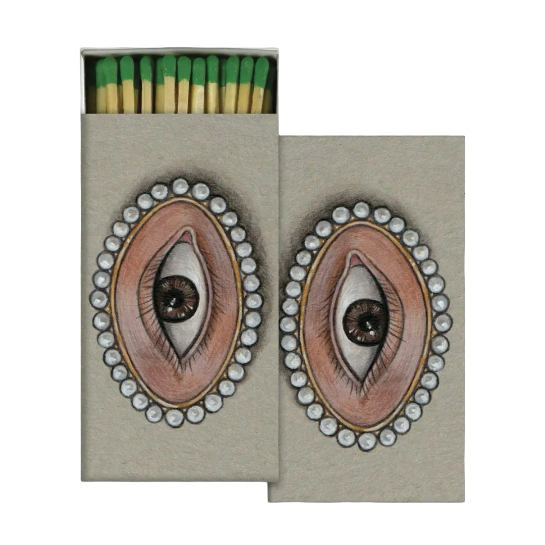 A decorative matchbox featuring an illustrated lover's eye portrait framed in a pearl bordered cameo on a gray background, shown open with green tipped matches inside.