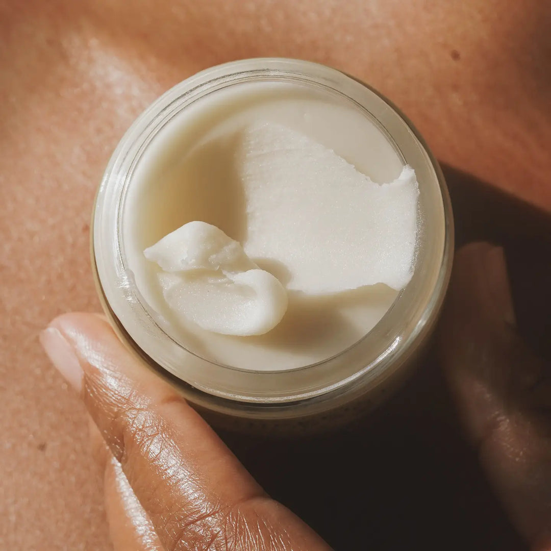 Enhanced view of creamy texture of deoderant in a glass jar held by a hand in front of bare skin. 